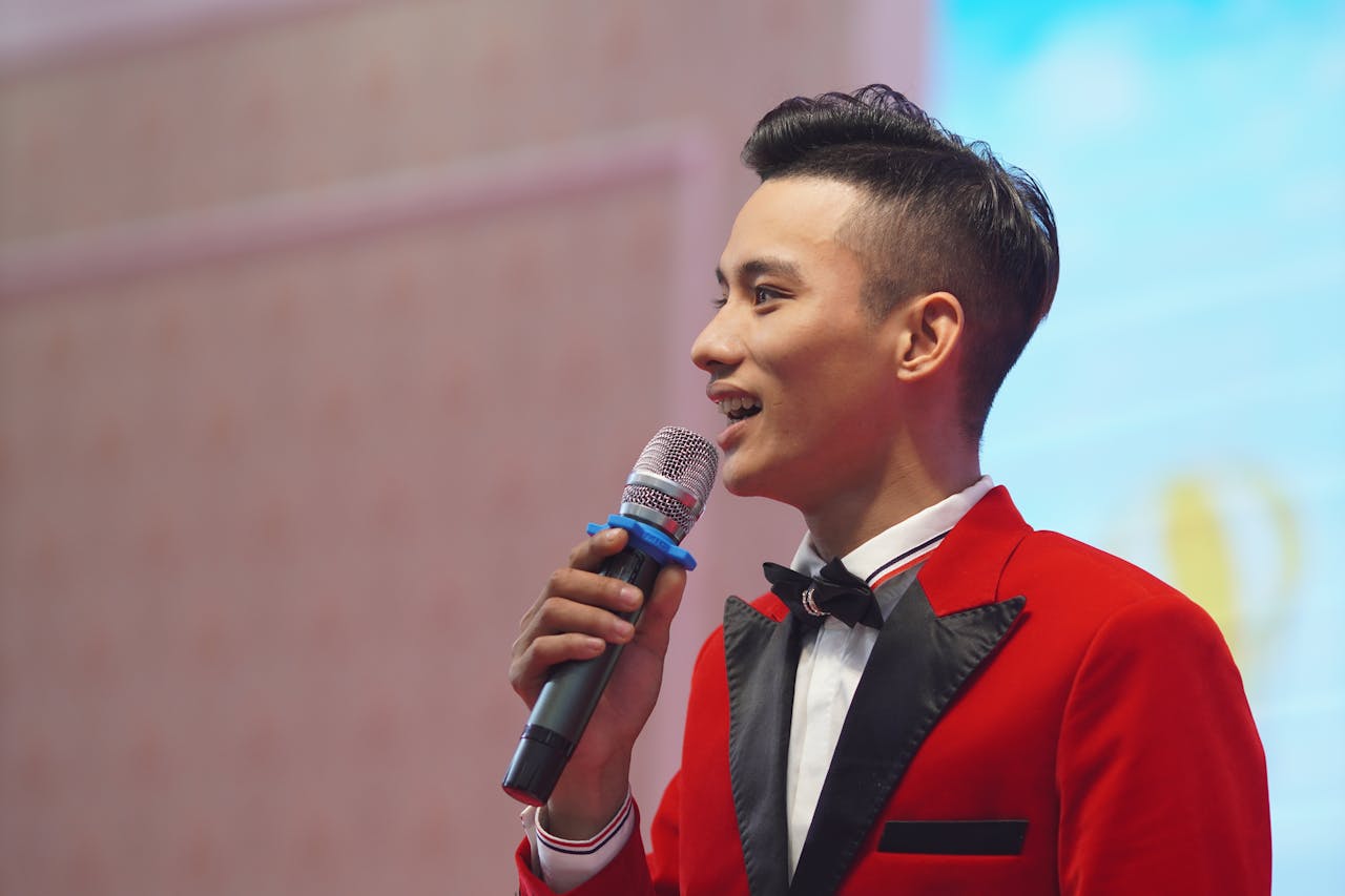 Asian male emcee in a stylish red suit holding a microphone, speaking at an indoor event.